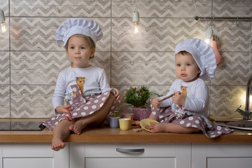 Two sisters one-year-old and three-years-old girls in a cook hats sit on the kitchen table and...