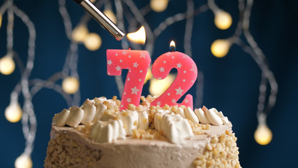Birthday cake with 72 number candle on blue backgraund set on fire by lighter. Close-up view