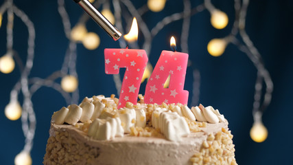 Birthday cake with 74 number candle on blue backgraund set on fire by lighter. Close-up view