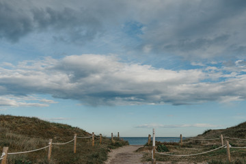 Fototapeta premium A walkway between sandy green dunes to the Mediterranean Sea in the afternoon. Clouds on the sky.
