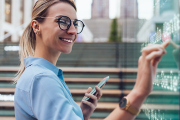 Interactive digital kiosk with public transport Info. Smiling female standing at big display with smartphone in hand.Young woman touching with finger interactive futuristic screen. Advanced technology