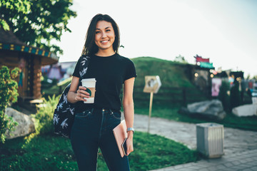 Half length portrait of beautiful asian hipster girl standing in park with coffee to go enjoying free time in summer, happy cheerful female student in casual trendy dress looking at camera in garden .