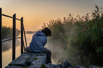 Junge sitzt träumend, bei Sonnenaufgang am Wasser
