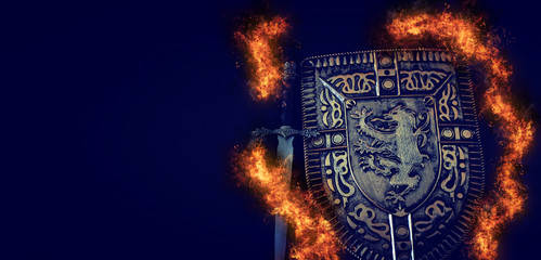 photo of shield knight armor and sword in fire flames over dark background. Medieval period concept