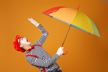 Sad mime man looking up with multi-colored umbrella in hand isolated on blank orange background
