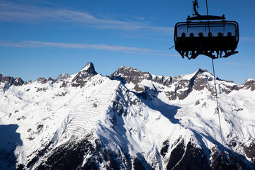 Silhouettes of people with skis and snowboards on a chairlift against a mountain panorama on a...