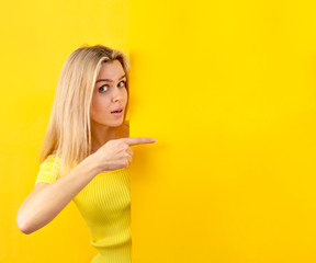 Young woman looking at camera with pointing gesture on a yellow wall. Copy space for banner