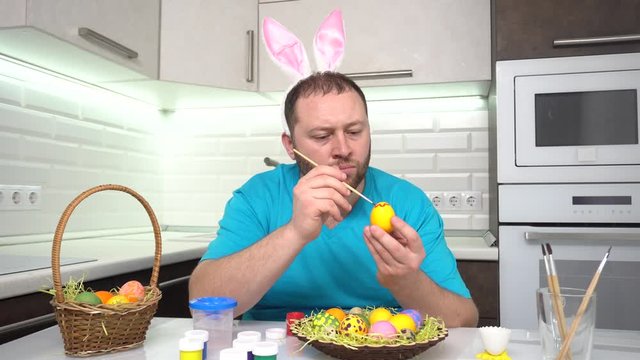 Happy Easter. A man in a rabbit ears blue t-shirt in the kitchen paints eggs preparing for Easter.