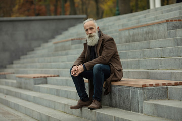 Portrait of a bearded senior man outdoors, sitting on a bench in a park.