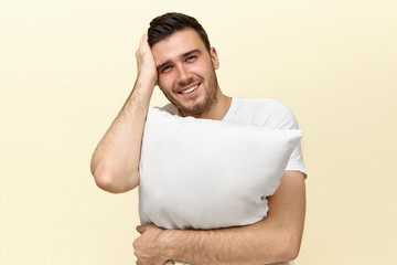 Studio image of handsome tired young Caucasian man looking at camera with happy smile needs some sleep because of stressful day at work, holding white orthopedic pillow, keeping hand on face