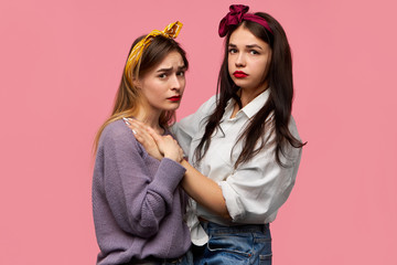 Portrait of two sad attractive stylish European female friends wearing stylish clothes and accessories posing isolated embracing each other, looking at camera with unhappy frustrated facial expression