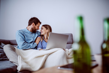 Cheerful cute caucasian couple sitting on sofa covered with blanket and using laptop for internet surf. In foreground are beer bottles. Living room interior.