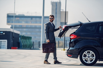 Full length of handsome caucasian businessman in suit and with sunglasses holding briefcase and closing trunk. Parking lot exterior.