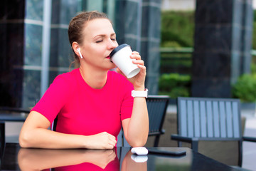 Beautiful happy relaxed girl, young pretty woman relaxing, enjoying coffee, dreaming outdoors on terrace and listening to music in wireless earphones with her eyes closed, red T-shirt, phone, gadget