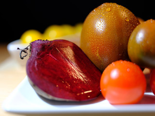 close up of variants of tomatoes and raw beetroot, covered with fresh water drops, on white tableware, in front of a dark background