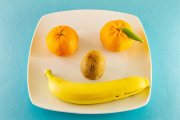 Tangerines kiwi and one banana on a white plate as smiling human face
