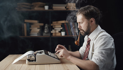 Portrait of focused bearded middle-aged male typing retro typewriter and smoking pipe, side view