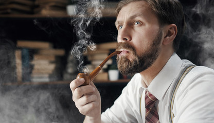 Portrait of thoughtful bearded mature man smoking wooden pipe over bookshelf background