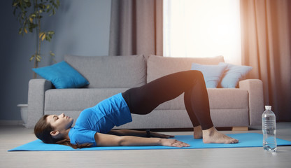 Young lady doing bridge pose training on yoga mat on floor in apartment