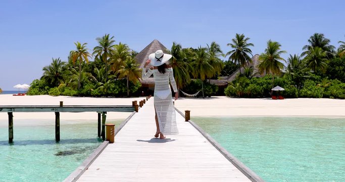 Eine attraktive Frau in wei&szlig;em Kleid und mit Hut l&auml;uft auf einem Steg auf eine tropische Paradies Insel mit t&uuml;rkisem Meer und Kokosnusspalmen zu