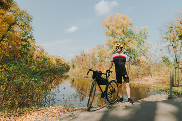 Obraz premium Portrait of man with bicycle standing on background of autumn landscape with river and looking to the side. Cyclist is resting on a bicycle by the lake. Cycling. concept