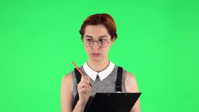 Portrait of funny girl in round glasses is angrily writing plans at black folder with pencil. Green screen