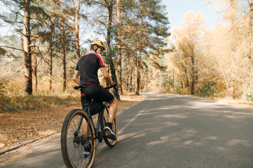 Obraz premium Back of a cyclist man in a sport bike suit and helmet stands on the road in the forest and looks away against the backdrop of an autumn landscape. Man rides a bicycle in a warm autumn day