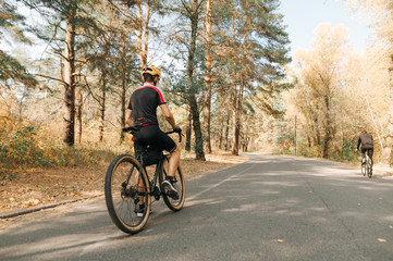 Naklejka premium Cyclist's back stands on the road in the forest on a background of autumn landscape. A man in a helmet and cycling clothes sits on a bicycle and rests after a ride. Weekend bicycle hobby.