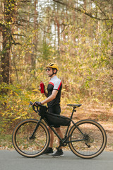 Obraz na płótnie Canvas Portrait of cyclist walking with bicycle and red water bottle on road in forest and looking away on background of autumn trees. The cyclist rests after the ride, drinks water and stands by the bike
