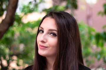 Portrait of a young brunette woman smiling. Outside sunny day with green leaves in the background.