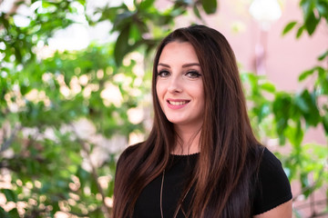 Portrait of a young brunette woman smiling. Outside sunny day with green leaves in the background.