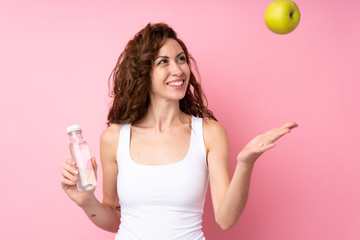 Young woman with curly hair holding an apple and a bottle of water over isolated pink background