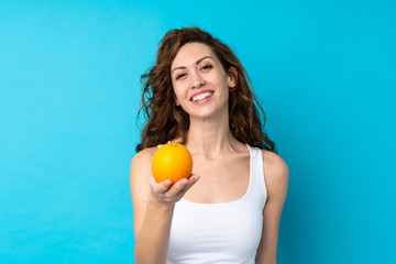 Young woman with curly hair holding an orange over isolated blue background