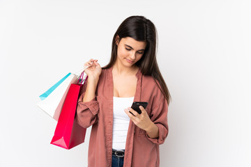 Young woman over isolated white background holding shopping bags and writing a message with her cell phone to a friend