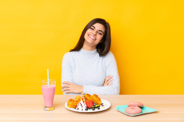 Young woman eating waffles and milkshake in a table over isolated yellow background laughing