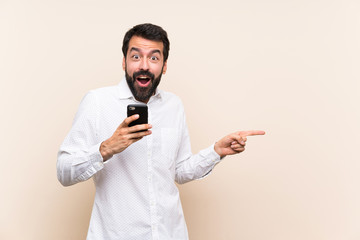 Young man with beard holding a mobile surprised and pointing finger to the side