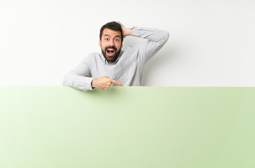 Young handsome man with beard holding a big green empty placard surprised and pointing finger to the side