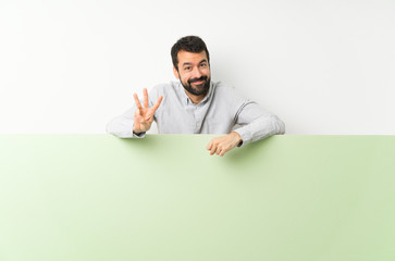 Young handsome man with beard holding a big green empty placard happy and counting three with...