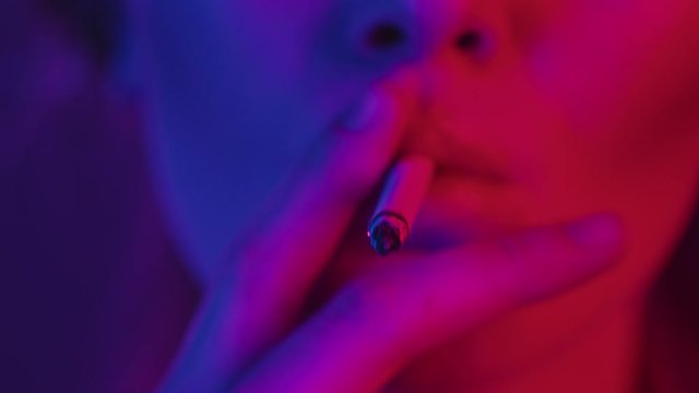 Woman lips sexually smoking cigarette focused closeup in neon lighting, slowmotion. Addicted sexy girl inhales smoke at nightclub underground party. Nightlife, alternative lifestyle concept