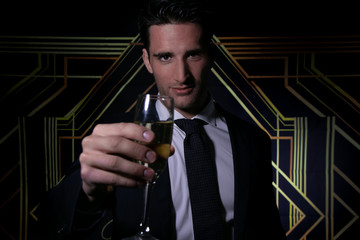 Handsome man in suit and tie in front of art deco backdrop holding a glass of sparkling wine to...