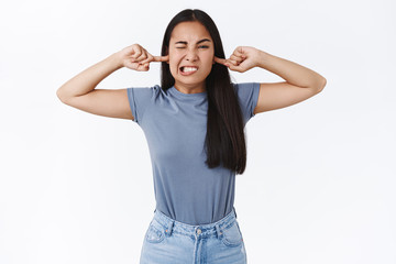 I am not hearing you. Silly and childish cute asian girl refuse listen to you as cover ears with fingers, cringe, squinting and clench teeth from bothering, disturbing loud noise, white background