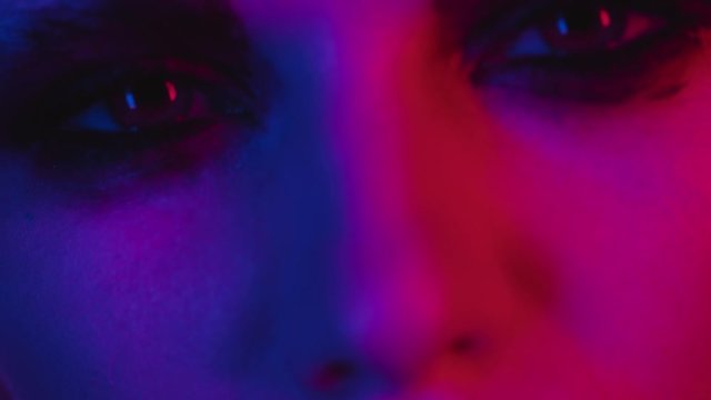 Fashion model portrait in colored neon light with dark makeup expressively looks into camera. Extreme closeup of eyes and lips. Provocative handsome girl focused shot