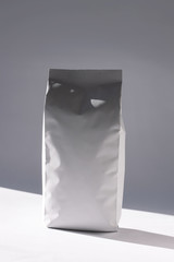 Blank white foil bag on monochrome background in minimal style with natural light shadow. Packaging for foods or drinks with valve and seal template mockup. Metallic coffee tea retail package design.