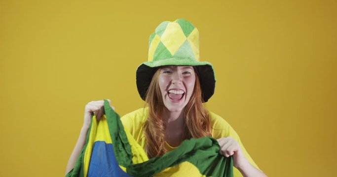 Brazil supporter. Brazilian redhead woman fan celebrating on soccer, football match on yellow background. Brazil colors. Wearing a t-shirt, flag and fan hat. 4K.