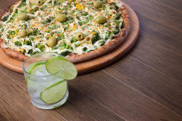 Broccoli pizza served on wooden board. Baked with mozzarella, broccoli, corn, peas and olives. Glass with carbonated water and lemon. Vegetarian. Horizontal photo, upper left alignment.