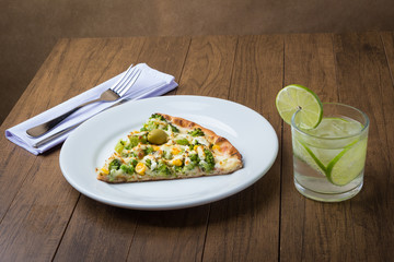 Broccoli pizza served on wooden board. Baked with mozzarella, broccoli, corn, peas and olives. Slice of pizza served on a plate. Soda water with sliced lemon. Horizontal photo.