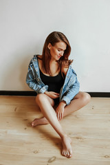 Stylish young girl during pregnancy sitting on a wooden floor posing in a denim jacket. Happy woman holds tummy with hand