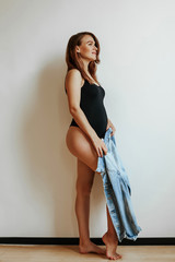 A pregnant girl with a slender figure holds a denim jacket in her hands and poses against the white wall in the studio. A woman with a slender figure in a black body.