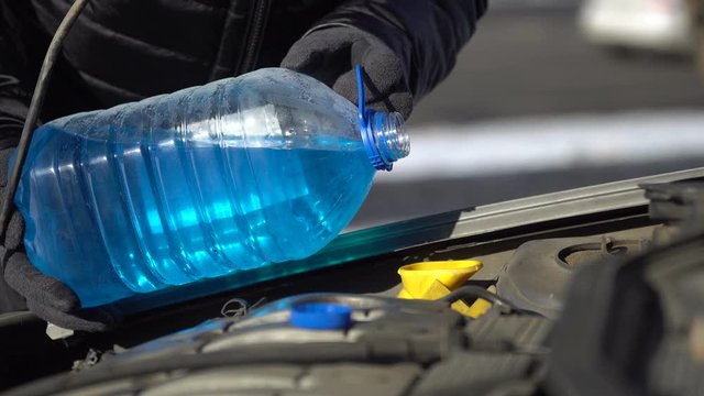 Male hands wearing winter gloves pouring an antifreeze liquid into a windshield washer tank of a car standing on the roadside. 4K resolution video.