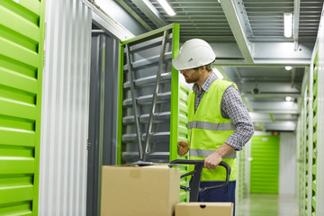 Manual worker in protective workwear standing with cart and loading boxes in storage room